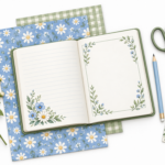 An open journal with floral pages rests on blue daisy and sage gingham papers, beside scissors, a blue pencil, a decorated tag, and white daisies.